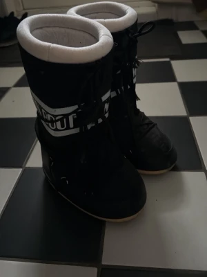 Svarta Moon Boots med snörning - Säljer ett par klassiska svarta Moon Boots med vit vadderad kant och snörning framtill. Skorna har en hög siluett och bred sula, perfekta för kalla dagar. Tydlig logga runt skaftet och mjukt foder för extra komfort. Storlek 38 men passar även 37 och 39 också!