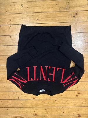 Svart tröja från Valentino med röd logga - Svart långärmad tröja från Valentino i 100% bomull. Tröjan har en stor röd Valentino-logga över bröstet och ärmarna, samt ribbade muddar vid hals, ärmslut och nederkant. Snygg och stilren design som sticker ut med den röda texten.