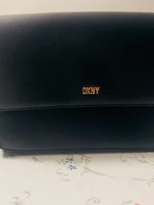 Svart axelväska från DKNY i skinn - Snygg svart axelväska från DKNY med stilren design och guldfärgad logga framtill. Väskan är tillverkad i slätt skinn och har en klassisk rektangulär form med lock. Perfekt storlek för att få plats med det viktigaste och passar till många olika outfits.