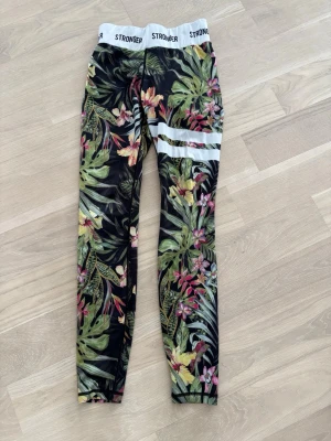 Blommiga leggings från Stronger - Snygga leggings från Stronger med tropiskt blommigt mönster i grönt, rosa och gult på svart botten. De har bred resår med logga upptill och två vita ränder på ena benet. Perfekta för träning eller chill.