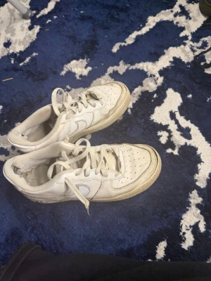 Vita Nike Air Force 1 sneakers - Säljer ett par klassiska vita Nike Air Force 1 sneakers med snörning och rund tå. Skorna har perforerade detaljer på tån och är tillverkade i skinn med en platt sula. Perfekta för dig som gillar streetwear och vill ha en ikonisk sneaker i garderoben.