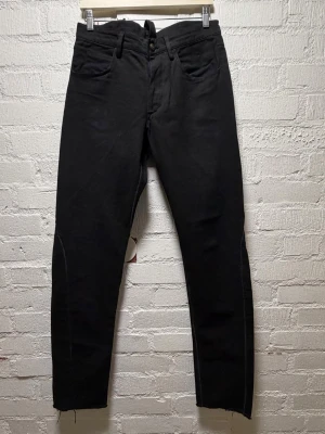 Zam Barrett Canvas Jeans - Svarta byxor med tapered passform och snygga sömdetaljer längs benen. Materialet är kanvas med en grov struktur. Byxorna har metallknappar med unika graveringar och råa kanter vid bensluten för en edgy vibe. Fickor både fram och bak. Mått: Midja 42 cm