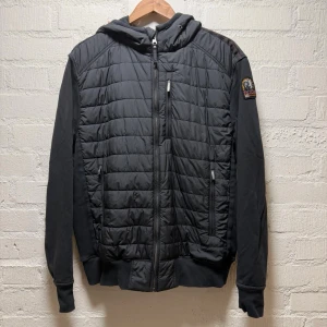 Parajumpers hybridjacka - Svart hybridjacka från Parajumpers med quiltad framsida, huva och snyggt märke på ärmen. Jackan har dragkedja framtill, två sidofickor med dragkedja och ribbade muddar. Perfekt för dig som gillar stilren och sportig look. Saknar snöre till luvan. I övrigt mycket fint skick. Liten i storleken.