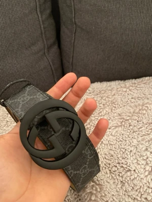 Gucceee bälte - Säljer ett svart bälte  med klassiskt GG-mönster över hela remmen och en stor, rund svart spänne med GG-logga. Bältet har en stilren look och är tillverkat i slitstarkt syntetmaterial med en matt finish. Perfekt för att lyfta din outfit.