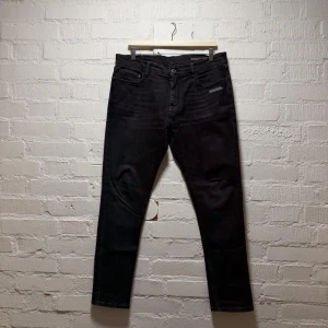 Off-White jeans - Säljer ett par svarta jeans från Off-White med vit diagonal print och text på bakfickan. Klassisk femficksmodell med dragkedja framtill och cool detalj med stor zip puller. Jeansen har en slim fit passform och är tillverkade i slitstark bomullsdenim. Saknar en av nitarna på fickorna, i övrigt mycket fint skick. 