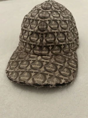 Brun mönstrad keps från Salvatore Ferragamo - Säljer en brun keps med ett snyggt, grafiskt mönster från Salvatore Ferragamo. Kepsen har böjd skärm och är tillverkad i ett slitstarkt bomullsmaterial. Perfekt för dig som vill sticka ut med en unik accessoar.