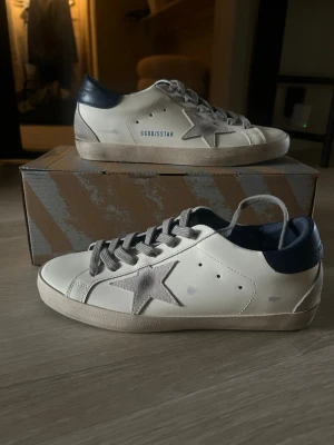 Golden Goose vita och marinblå sneakers - Golden Goose sneakers i vitt skinn med marinblå häl och grå mockadetaljer. Klassisk stjärna på sidan, grå snörning och GGDB/SSTAR-tryck. Snygg kontrast mellan material och färger, perfekt för dig som gillar streetstyle med premiumkänsla.
