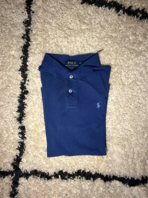 Blå pikétröja från Polo Ralph Lauren - Snygg blå pikétröja från Polo Ralph Lauren med klassisk krage och två knappar framtill. Tröjan har en broderad logga på bröstet och är gjord i mjuk bomull. Skriv om funderingar eller om du vill ha fler bilder.  Slim fit-modell som sitter schysst på kroppen. Storlek 14-16/ XS 
