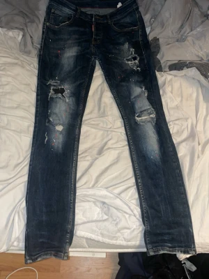 Dsquared2 blå slitna jeans straight fit - Säljer ett par blå jeans från Dsquared2 med straight fit. Jeansen har slitningar och hål på benen samt färgstänk för en edgy look. Klassisk femficksmodell med Dsquared2-logga på röd etikett och bakficka. Tillverkade i jeansmaterial med kontrastsömmar.