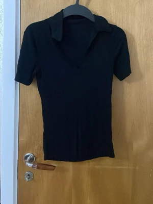 Svart polotopp med krage - En svart polotopp med korta ärmar och klassisk krage. Toppen har en stilren och enkel design utan mönster, perfekt att matcha med olika outfits. Materialet känns mjukt och stretchigt, vilket gör den bekväm att bära hela dagen.