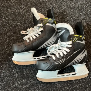 Svarta skridskor från CCM - Säljer ett par svarta skridskor från CCM med vita snören och grå detaljer. De har en robust design med vadderad insida för extra komfort. Perfekta för isaktiviteter och har en stilren look.