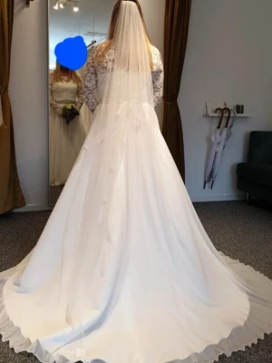 Brudklänning - Säljer en vit brudklänning med vackra spetsdetaljer på överdelen och långa ärmar. Kjolen är vid och svepande med volangkant. Midjan pryds av ett glittrigt strassbälte som ger extra lyxig känsla. Går att ta av. Perfekt för dig som söker en vintage feeling på din bröllopsdag. 