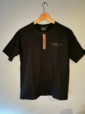 Svart Essentials Fear of God t-shirt - Säljer en svart t-shirt från Essentials Fear of God med tryck fram och bak. Klassisk rund hals och korta ärmar. Tillverkad i mjuk bomull för skön känsla och clean look. Perfekt för dig som gillar streetwear och vill ha en enkel men snygg tee.