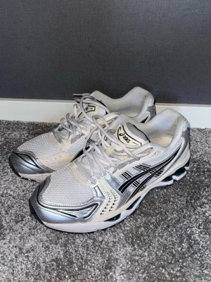 Asics Gel Kayano 14 - Säljer ett par snygga Asics sneakers i storlek 43.5 med silvriga och vita detaljer samt svarta ränder på sidorna. Skorna har mesh-ovandel för bra ventilation och snörning framtill. Sulan är robust och har svart och vit mönstring för extra grepp. Bra skick
