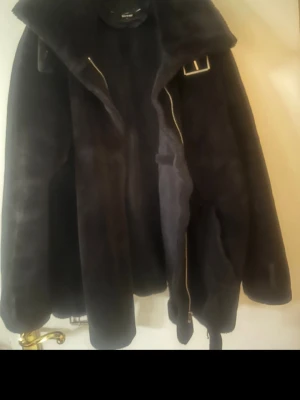 Svart teddyjacka - Mysig svart teddyjacka med bred krage, dragkedja och bälte i midjan. Jackan har detaljer i metall, som spännen och öglor, samt långa ärmar. Perfekt för kalla dagar och ger en cool look till vilken outfit som helst.