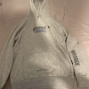 Grå hoodie från Hoodrich xs  - Säljer en grå hoodie från Hoodrich i storlek XS. Tröjan har tryck både fram och bak med texten HOODRICH i svart och blått. Klassisk huva med snörning och känguruficka framtill. Perfekt för chill dagar och streetstyle.