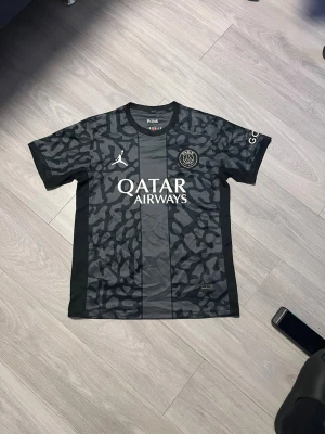 Svart PSG x Jordan fotbollströja - Säljer en svart PSG x Jordan fotbollströja med grått camouflage-mönster och vita loggor på bröstet. Tröjan har rund hals och korta ärmar, med Qatar Airways tryckt framtill. Perfekt för dig som gillar streetwear och fotboll.