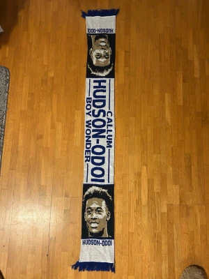 Blå och vit fotbollshalsduk Hudson-Odoi - Chelsea FC - Säljer en blå och vit fotbollshalsduk med texten 'Callum Hudson-Odoi Boy Wonder' och porträtt på båda sidor. Halsduken har fransar i ändarna och är perfekt för att visa support på matchen eller i vardagen. Chelsea FC