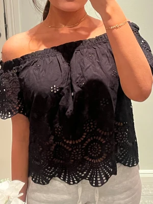 Svart offshoulder blus med spets - Säljer en svart offshoulder blus med vackra broderade spetsdetaljer och hålmönster. Blusen har korta ärmar och en böljande kant nedtill som ger en cool och feminin vibe. Perfekt för dig som gillar att sticka ut med detaljer och luftig stil.