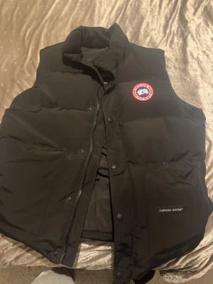 Svart dunväst från Canada Goose L/XL - Svart dunväst från Canada Goose i storlek XL sitter mer som L . Västen har en hög krage, klassisk logga på bröstet och dragkedja framtill med tryckknappar. Materialet är slitstark polyester och dunfyllning som håller dig varm. Perfekt för kalla dagar och snygg att styla över hoodie. 