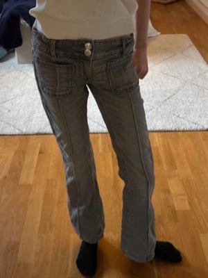 Grå bootcut jeans med dubbla knappar - Säljer ett par grå jeans med bootcut passform och dubbla knappar i midjan. Byxorna har stora framfickor och markerade sömmar längs benen för en cool vintage vibe. Materialet är klassisk jeans och färgen är en snygg tvättad grå. Från gina tricot young storlek 140.