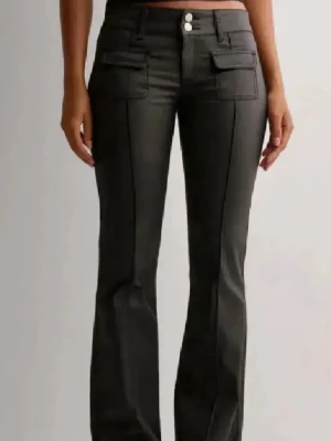 Svarta bootcut byxor med fickor - Snygga svarta bootcut byxor med två framfickor med lock och dubbla knappar i midjan. Byxorna har en markerad söm längs benen och är tillverkade i ett material som ger en stilren look. Perfekta för dig som vill ha en cool och trendig siluett.