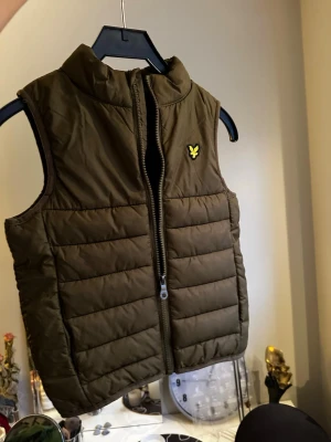 Brun dunväst från Lyle & Scott Junior - Säljer en brun dunväst från Lyle & Scott Junior med gul logga på bröstet. Västen har quiltad design, hög krage och dragkedja framtill. Perfekt för lager-på-lager och enkel att matcha med hoodies eller tröjor. Snygg och praktisk för kyliga dagar.
