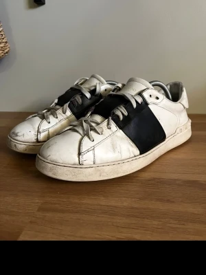 Valentino Garavani vita sneakers - strl.44 - Säljer ett par vita Valentino Garavani sneakers med bred svart läderdetalj över mitten. Skorna har rund tå, vita snören och platt sula. Klassisk logga på plösen och diskreta nitar bak på hälen. Tillverkade i skinn för en lyxig känsla. Se bilder för skick.