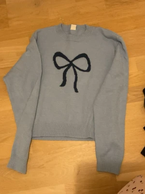 Ljusblå stickad tröja med rosett Lindex - Supersöt ljusblå stickad tröja från Lindex med en stor mörkblå rosett på bröstet. Tröjan har långa ärmar, ribbade muddar och rund hals. Perfekt för dig som gillar gulliga detaljer och vill ha något mysigt till garderoben.