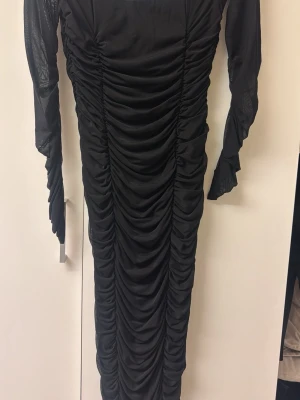 Svart långklänning med rynkade detaljer - Säljer en svart långklänning med rynkade detaljer längs hela klänningen. Klänningen har långa ärmar i mesh och en figurnära passform. Perfekt för dig som vill ha en stilren och trendig look.