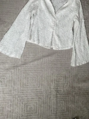 Vit blus med vida ärmar och spets - Säljer en vit blus med vida långärmar och snygg spetsdetalj över hela tyget. Blusen har en lätt croppad passform och knappar framtill. Perfekt för dig som gillar romantisk och trendig stil.