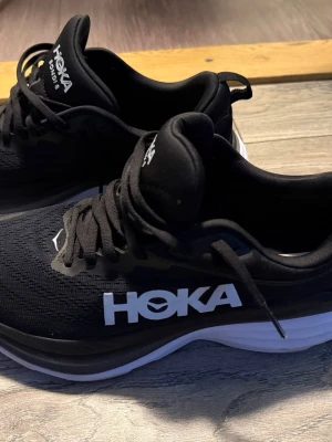 Svarta Hoka Bondi 8 sneakers - Säljer ett par svarta Hoka Bondi 8 sneakers med vit sula och tydlig Hoka-logga på sidan. Skorna har snörning och en rund tå, samt en tjock och mjuk plattformssula som ger grym komfort. Ovandelen är i syntetmaterial med mesh för bra ventilation.