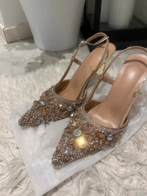 Glittriga pumps med spetsig tå - Säljer ett par glittriga pumps med spetsig tå och öppen häl. Skorna är dekorerade med stora pärlor och strass i blommönster, har en champagnefärgad ton och smala remmar med spänne. Klacken är smal och i metallic. Perfekta för att sticka ut med extra bling.