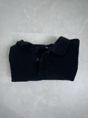 Svart kortärmad pike - Säljer en svart pike med klassisk krage och knappar framtill. Toppen har korta ärmar och är tillverkad i mjukt bomullsmaterial. Enkel och stilren design som funkar till många olika outfits.