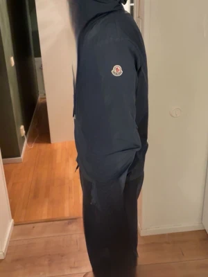 Mörkblå vindjacka från Moncler - Snygg mörkblå vindjacka från Moncler med huva och logotyp på ärmen. Jackan har dragkedja framtill och diskreta snörningar vid midjan. På huvan finns en cool randig detalj i rött och vitt. Perfekt för dig som gillar stilren design och premiumkänsla.