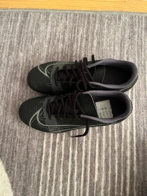 Nike Mercurial svarta fotbollsskor - Säljer ett par svarta Nike Mercurial fotbollsskor med silvergrå detaljer och snörning. Skorna har en rund tå och platt sula med dobbar för gräsplan. Ovandelen är tillverkad i syntetmaterial med ett grafiskt mönster och Nike-logga på sidan. Perfekt för dig som vill ha stil på planen.
