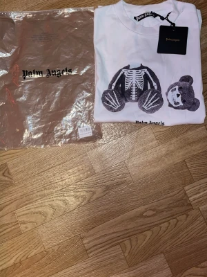 Vit Palm Angels t-shirt med björntryck - Säljer en vit t-shirt från Palm Angels med ikoniskt björntryck där huvudet är avskilt från kroppen. T-shirten har rund hals och korta ärmar, samt Palm Angels-logga tryckt på bröstet. Materialet är mjuk bomull och passformen är normal.