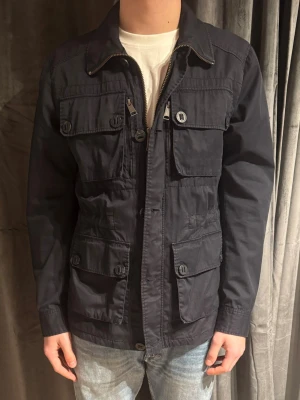 Mörkblå vindjacka från J.Lindeberg - Säljer en svart vindjacka från J.Lindeberg i storlek M. Jackan har flera stora fickor med knappar, dragkedja framtill och klassisk krage. Materialet är lätt och smidigt, perfekt för vår och höst. Enkel och stilren design som funkar till det mesta. Pris kan diskuteras vid snabb affär 