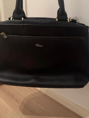 Svart handväska från Don Donna - Snygg svart handväska från Don Donna i strukturerat skinnimitation. Väskan har guldfärgade detaljer, flera fack med dragkedja och ljusbeige innerfoder. Perfekt storlek för att få plats med allt du behöver och stilren design som passar till det mesta.