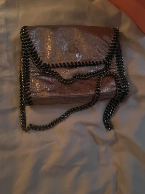 Glittrig silver axelväska med kedja - Säljer en glittrig silverfärgad axelväska med kraftig kedja i metall och snygga detaljer längs kanterna. Väskan har ett fack med magnetlås och plats för mobil och småsaker. Perfekt när du vill sticka ut lite extra.