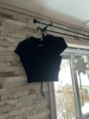 Svart croppad ribbad topp med snörning - Säljer en svart croppad topp  med ribbad struktur och snygg snörning framtill. Toppen har korta ärmar och v-ringad hals. Perfekt att styla med jeans eller kjol för en cool look.