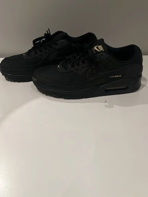Nike Air Max 90 helsvarta sneakers - Säljer ett par Nike Air Max 90 i helsvart med diskreta gulddetaljer. Skorna har klassisk chunky sula med synlig Air-enhet, snörning och ovandel i syntet och skinn. Perfekta för dig som gillar stilrena och sportiga sneakers med retrovibe.