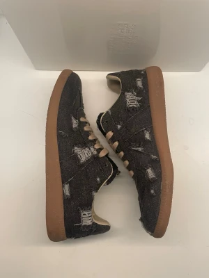 Unika Maison Margiela Skor  - Unika Maison Margiela Replica sneakers i svart denim med distressed detaljer och beige snörning. Skorna har en klassisk gummisula i brunt och insida i ljusbeige. 