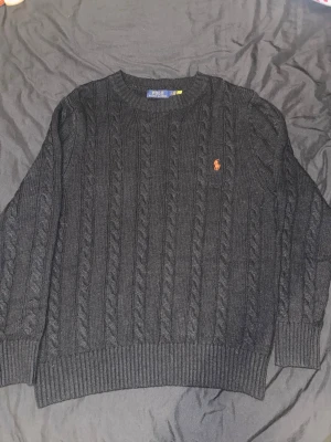 Svart kabelstickad tröja Polo Ralph Lauren - Kabelstickad svart tröja från Polo Ralph Lauren med klassisk rund halsringning och broderad logga i orange på bröstet. Tröjan har ribbade muddar och är långärmad, perfekt för kyligare dagar.