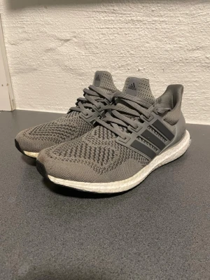 Grå Adidas Ultraboost sneakers - Säljer ett par grå Adidas Ultraboost sneakers med stickad ovandel och klassiska tre ränder på sidan. Skorna har snörning, vit boost-sula och en modern, sportig look. Perfekta för dig som gillar stilrena och bekväma sneakers.