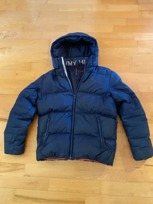 Blå pufferjacka från Tommy Hilfiger - Säljer en mörkblå pufferjacka från Tommy Hilfiger med huva och dragkedja framtill. Jackan har två sidofickor med dragkedja och diskret logga på bröstet. Perfekt för kalla dagar och riktigt skön att bära.