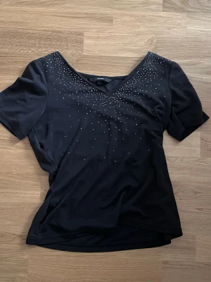 Svart topp med paljetter Vero Moda XS - Snygg svart topp från Vero Moda i storlek XS. Toppen har korta ärmar, v-ringning och är dekorerad med små silvriga paljetter över bröstet. Tillverkad i mjukt material som känns skönt mot huden. Perfekt för dig som vill ha något enkelt men ändå glittrigt. Har sytt om i midjan så det blir lite utsvängt där nere!