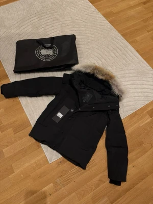 Svart dunjacka från Canada Goose - Säljer en svart dunjacka från Canada Goose med avtagbar huva och snygg pälskant. Jackan har flera fickor, dragkedja och knappar framtill samt klassisk patch på ärmen. Perfekt för kalla dagar och riktigt varm. Stilsäker och tidlös design.