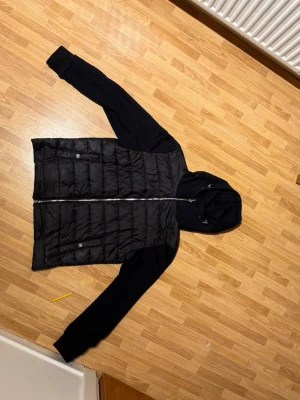 Svart pufferjacka med huva från H&M - Säljer en svart pufferjacka från H&M med quiltad framsida, mjuka ärmar och stor huva. Jackan har dragkedja framtill, två fickor med knapp och en clean look. Perfekt för dig som gillar streetwear och vill ha något enkelt men snyggt.