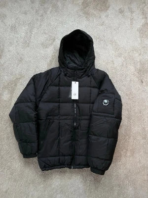 C.P. Company Jacka  - Svart pufferjacka från C.P. Company med huva och ikonisk linsdetalj på ärmen. Stilren och perfekt till Svensk vinter. Helt ny och oanvänd. 